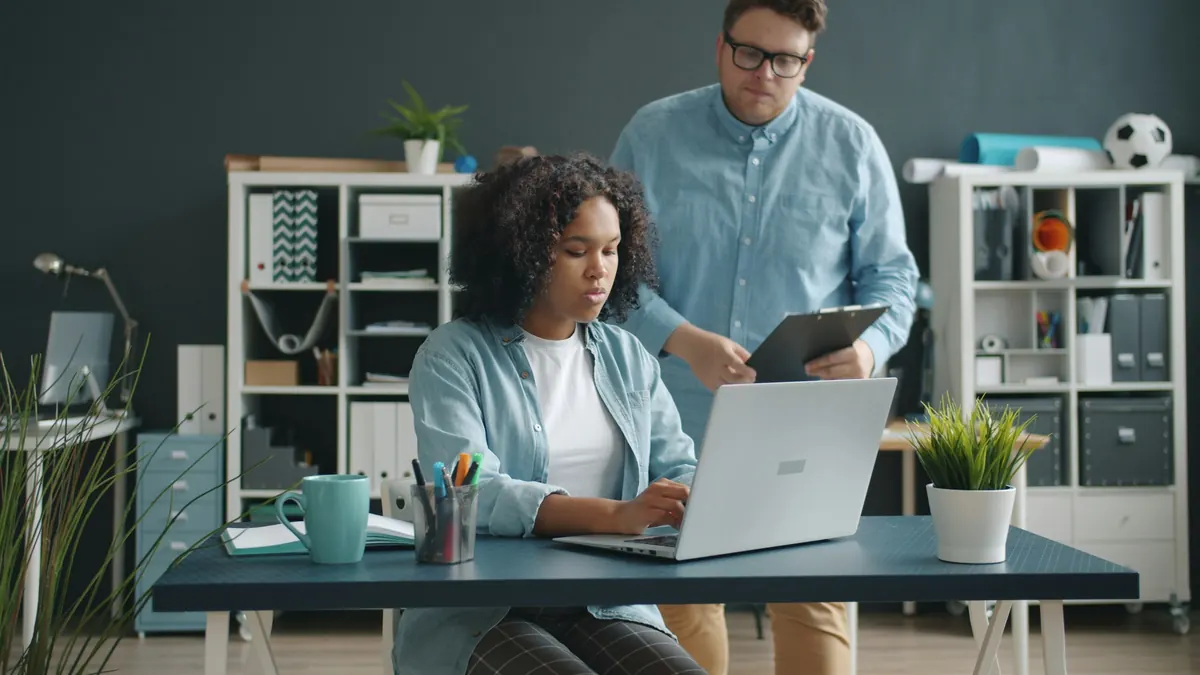 Team member working on a laptop while a colleague reviews notes on a clipboard